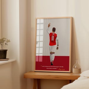 May include: A framed print featuring a stylized illustration of a soccer player in a red and white jersey with the number 11. The print includes a quote in white text on a red background. The frame is light brown.