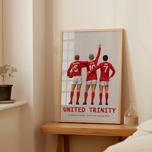 Op de afbeelding: Een ingelijste print met drie voetballers in rode shirts en witte shorts. De print leest "UNITED TRINITY" met de namen Sir Bobby Charlton, Denis Law en George Best. Eén speler heeft zijn arm omhoog.