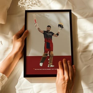 May include: Framed art print featuring a cricket player in a black and red uniform, holding a bat and helmet. The artwork has a minimalist style with a quote at the bottom. The background is a mix of beige and red.