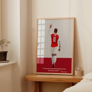 May include: A framed print of a soccer player wearing a red and white jersey with the number 11 on the back. The player is raising his right hand in the air. The quote "For me, scoring goals and providing assists is the same. They both win you matches." - Mesut Özil is printed below the image.