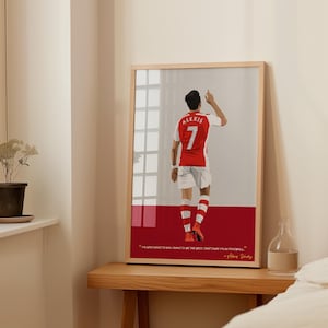 May include: A framed illustration of a soccer player wearing a red and white jersey with the number 7 on the back. The player is facing away from the viewer and has his arm raised in the air. The quote "I always want to win, I want to be the best, that's why I play football." is printed below the image. The quote is attributed to Alexis Sanchez.