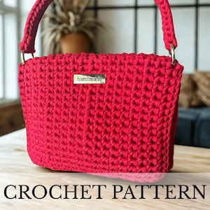Op de afbeelding: Een handgemaakte, rode gehaakte handtas met een bijpassende handgreep en zilverkleurige hardware. De tas heeft een gestructureerd steekpatroon en een klein metalen label met de tekst "handmade". De woorden "CROCHET PATTERN" worden onderaan de afbeelding weergegeven.