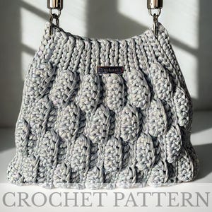 May include: A light gray crocheted handbag with a textured, bubble-like pattern. The bag has silver-toned hardware and a braided handle. The words "CROCHET PATTERN" are printed at the bottom of the image. The bag is handmade.