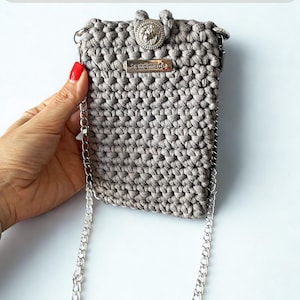 Può includere: Una borsa a tracolla in uncinetto grigio con tracolla a catena in metallo argentato, chiusura a bottone circolare con emblema a testa di leone e una piccola targhetta metallica rettangolare con la scritta "handmade". Testo "CROCHET PATTERN".