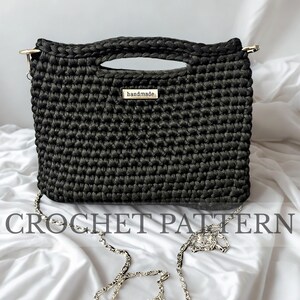 Può includere: Una borsa nera all'uncinetto con una tracolla a catena argentata e un manico superiore. La borsa ha una forma rettangolare e una piccola etichetta metallica con la scritta "handmade". Le parole "CROCHET PATTERN" sono stampate sotto la borsa.