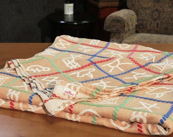 May include: A folded tan throw blanket with a woven pattern of white rope designs and red, blue, and green accents. The blanket has a "Patjes" label.