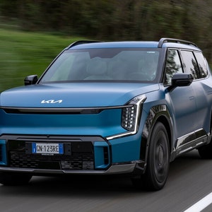 May include: A blue Kia EV6 electric SUV driving on a paved road with green trees in the background. The car has a license plate that reads "CN-123RE".