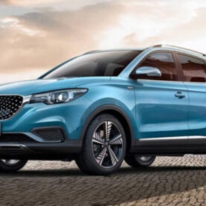 Puede incluir: Un SUV eléctrico azul claro con un cable de carga conectado a la parte delantera del coche. El coche tiene una parrilla negra con un logotipo MG plateado. La matrícula dice "MG 1 EZS 001".