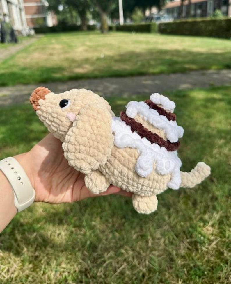 K&ouml;nnte beinhalten: Ein beiges H&auml;kel-Pl&uuml;schtier in Hundeform mit brauner Nase, schwarzen Augen und rosa Wangen. Auf dem R&uuml;cken des Hundes befindet sich ein wei&szlig;-braunes Zimtschnecken-Design mit strukturierten Garnschlingen und einem kleinen Schwanz am Ende.