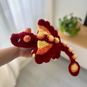 May include: A handmade crochet dragon in shades of red, orange, and yellow. The plush toy features a detailed design with wings, spikes, and a long tail. The dragon is held in a person's hand.