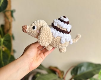 May include: A crocheted plush toy shaped like a dachshund dog with light beige yarn, featuring a dark brown nose, black eyes, and a cinnamon roll-inspired back with white and dark brown yarn swirls. The toy has floppy ears and a small tail.