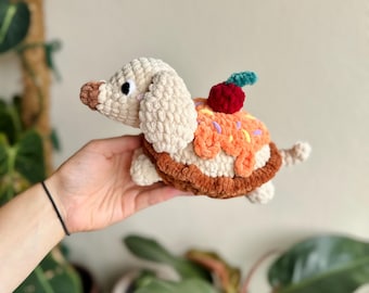 May include: A crocheted plush toy shaped like a dog with a tan body, floppy ears, and a brown nose. The dog has a shell on its back resembling a pastry with orange frosting, colorful sprinkles, and a small red cherry with a green stem on top.