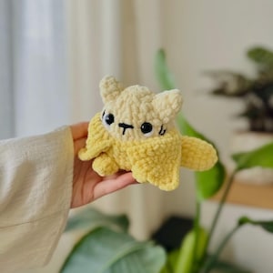Puede incluir: Un pequeño peluche de ganchillo hecho a mano con forma de gato que emerge de una cáscara de plátano amarilla. El gato es de color crema claro con grandes ojos de seguridad de plástico negro y una nariz y boca negras bordadas.