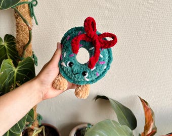 Könnte beinhalten: Ein gehäkeltes Plüsch-Donut-Spielzeug in Dunkelgrün mit einer leuchtend roten Samtschleife oben, kleinen schwarzen Sicherheitsaugen, einem gestickten Lächeln und rosa-weißen Garnstreuseln. Der Donut hat zwei kleine, hellbraune Plüschfüße an der Unterseite.