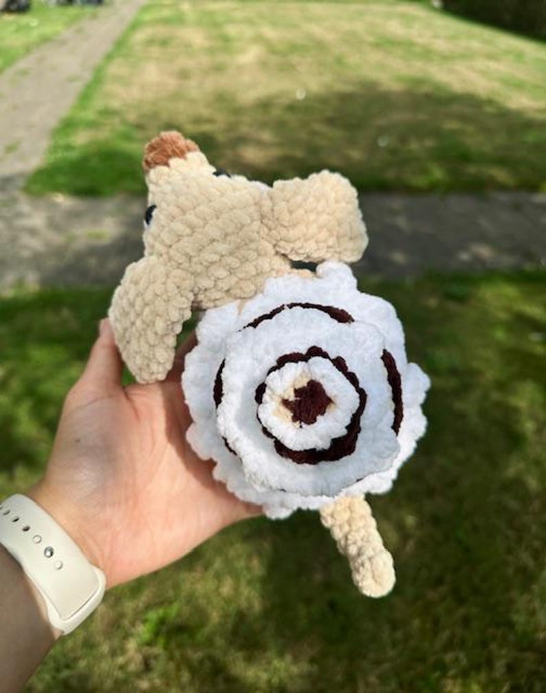 K&ouml;nnte beinhalten: Ein handgefertigtes H&auml;kel-Pl&uuml;schtier in Hundeform aus hellbraunem Garn, mit einem gro&szlig;en, spiralf&ouml;rmigen, wei&szlig;-dunkelbraunen blumenartigen Muster auf dem R&uuml;cken, einer kleinen braunen Nase und dunklen Augen aus weichem, strukturiertem Pl&uuml;schgarn.