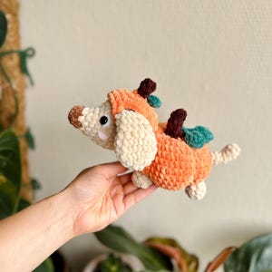 May include: A crochet plush toy shaped like a dog wearing a pumpkin costume. The dog has cream-colored yarn, black eyes, and pink cheeks. The pumpkin suit is orange with a brown stem and green leaves, featuring a textured, soft yarn finish.