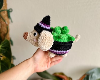 May include: A crocheted plush dog wearing a black, purple, and white pointed hat. The dog has a cream-colored body, a brown nose, and a black and purple saddle featuring a bright green, textured succulent plant attached to its back.