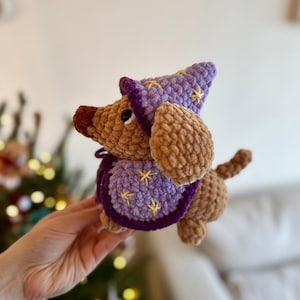 May include: A crocheted dachshund dog wearing a purple wizard hat and cape, adorned with yellow star accents. The dog has brown fur and floppy ears. The cape and hat are a darker shade of purple, creating a whimsical, handmade aesthetic.