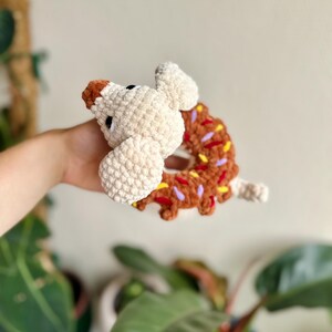 May include: A crocheted plush toy shaped like a brown donut with colorful yarn sprinkles, featuring a cream-colored dog head with a brown nose and black eyes attached to the side of the donut, crafted from soft, textured yarn material.