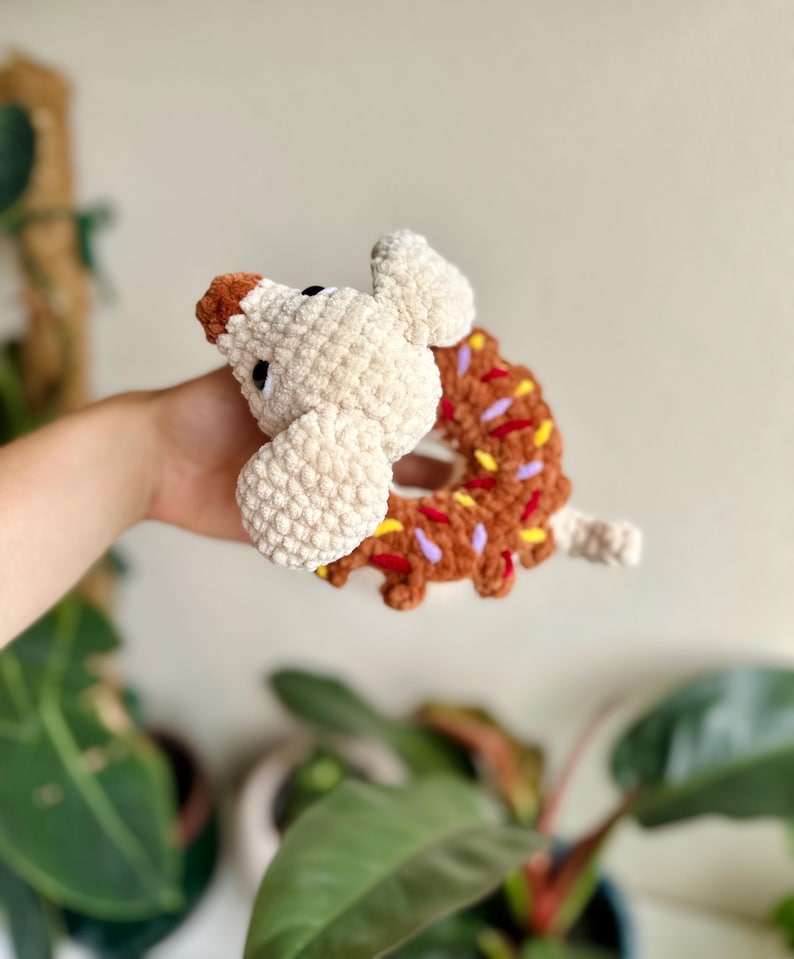 May include: A crocheted plush toy shaped like a brown donut with colorful yarn sprinkles, featuring a cream-colored dog head with a brown nose and black eyes attached to the side of the donut, crafted from soft, textured yarn material.