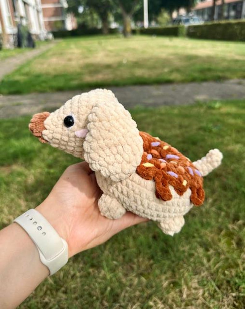 May include: A crocheted plush dog toy in a light beige color with a brown nose and black eyes. The back of the dog features a brown textured patch resembling icing with small, colorful sprinkles in yellow, orange, and purple hues.