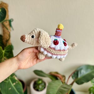 Pode incluir: Um brinquedo de crochê em formato de cão basset na cor bege, com um corpo de bolo branco decorado com cerejas vermelhas e uma vela listrada de amarelo e roxo nas costas. O cão tem nariz marrom, olhos pretos e bochechas rosadas.