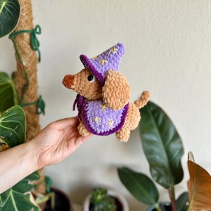 Könnte beinhalten: Ein gehäkelter Plüschhund in Beige, der einen violetten Zaubererhut und einen passenden Umhang trägt. Sowohl der Hut als auch der Umhang sind mit gestickten gelben Sternen verziert. Der Hund hat eine dunkelbraune Nase und schwarze Augen.