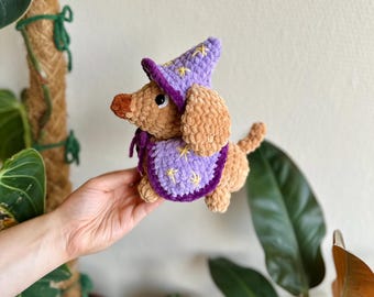 May include: A tan crocheted plush dog wearing a purple wizard hat and a matching purple cape. Both the hat and cape feature embroidered yellow star details. The dog has a dark brown nose and black plastic eyes, crafted from soft yarn.