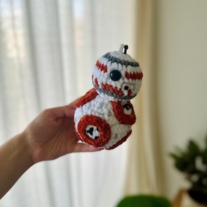 May include: A spherical crochet toy featuring a white, orange, and grey color scheme with a rounded head and body. The toy has black plastic eyes, embroidered details, and a small antenna on top, crafted with a soft, textured yarn finish.