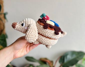 May include: A crocheted plush toy shaped like a dachshund with a tan body and floppy ears. The back of the dog features a stack of pancakes topped with dark brown syrup, a red strawberry, a yellow butter pat, and a blue blueberry accent.