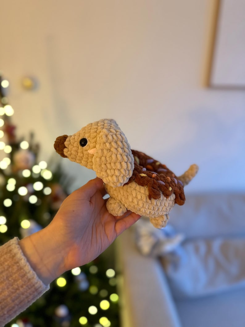 May include: A crocheted plush toy shaped like a dachshund dog with a tan body, a dark brown nose, and a dark brown donut-shaped back featuring small colorful sprinkles. The toy has black plastic eyes and a small pink stitched detail on the cheek.