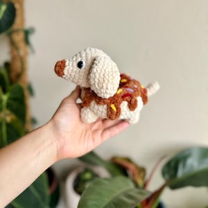 May include: A crochet plush toy shaped like a dachshund dog with a cream-colored body and a brown snout. The dog's back features a brown textured section resembling a donut with colorful yarn sprinkles in yellow, purple, and red.