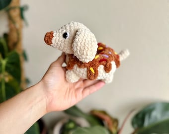 May include: A crochet plush toy shaped like a dachshund dog with a cream-colored body and a brown snout. The dog's back features a brown textured section resembling a donut with colorful yarn sprinkles in yellow, purple, and red.