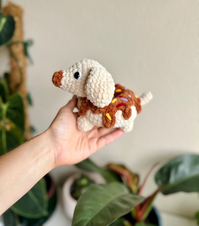 May include: A crochet plush toy shaped like a dachshund dog with a cream-colored body and a brown snout. The dog's back features a brown textured section resembling a donut with colorful yarn sprinkles in yellow, purple, and red.