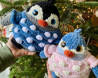 May include: Two plush penguin toys made of soft yarn. The larger penguin is black, white, and gray, wearing a blue sweater with white pom-poms. The smaller penguin is blue and white, wearing a light pink sweater with white pom-poms.