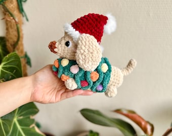 Könnte beinhalten: Ein beiges, gehäkeltes Dackel-Stofftier, das eine rote Weihnachtsmütze mit weißem Bommel und einen dunkelgrünen, gehäkelten Pullover mit kleinen, bunten Bommeln trägt. Das Stofftier hat schwarze Augen und eine braune Nase aus weichem Garn.