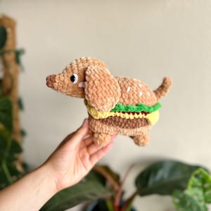 Pode incluir: Um brinquedo de pelúcia de crochê em forma de cão dachshund combinado com um hambúrguer. O cão tem pelo castanho claro, nariz castanho escuro e olhos pretos. O corpo apresenta camadas de alface verde, queijo amarelo e um hambúrguer.