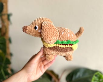 May include: A plush crochet toy shaped like a dachshund dog combined with a hamburger. The dog has light brown fur, a dark brown nose, and black eyes. Its body features layers of green lettuce, yellow cheese, and a brown burger patty.