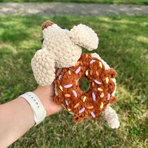 May include: A crocheted plush toy featuring a beige dog with floppy ears holding a brown donut with colorful sprinkles. The donut has a central hole and is textured with soft yarn, while the dog has a small brown nose and dark eyes.