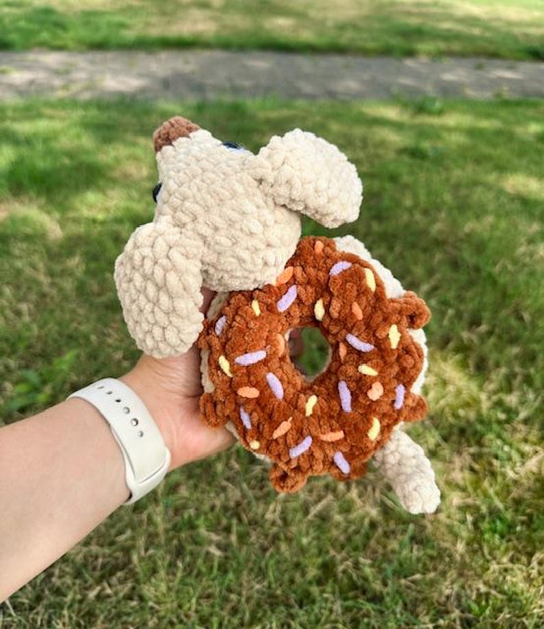 May include: A crocheted plush toy featuring a beige dog with floppy ears holding a brown donut with colorful sprinkles. The donut has a central hole and is textured with soft yarn, while the dog has a small brown nose and dark eyes.
