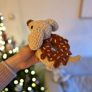 May include: A crochet plush toy shaped like a tan dog with floppy ears, featuring a dark brown donut with colorful sprinkles around its midsection. The toy is crafted from soft, textured yarn and includes a small tail and dark plastic eyes.