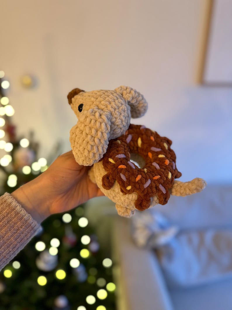 May include: A crochet plush toy shaped like a tan dog with floppy ears, featuring a dark brown donut with colorful sprinkles around its midsection. The toy is crafted from soft, textured yarn and includes a small tail and dark plastic eyes.