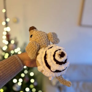 K&ouml;nnte beinhalten: Ein geh&auml;keltes Pl&uuml;schtier in Form einer Schnecke mit beigefarbenem K&ouml;rper und einem gro&szlig;en, spiralf&ouml;rmigen Geh&auml;use aus wei&szlig;em Garn mit dunkelbraunen Akzenten. Die Schnecke hat eine kleine braune Nase, dunkle Augen und eine weiche Textur.