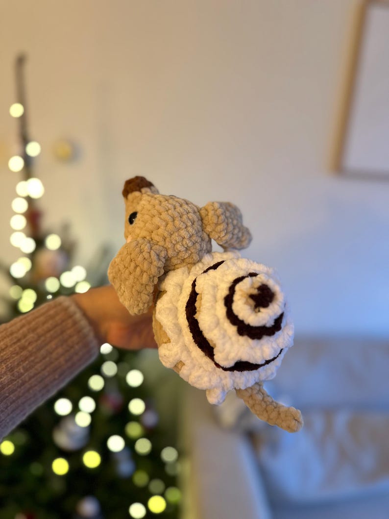 K&ouml;nnte beinhalten: Ein geh&auml;keltes Pl&uuml;schtier in Form einer Schnecke mit beigefarbenem K&ouml;rper und einem gro&szlig;en, spiralf&ouml;rmigen Geh&auml;use aus wei&szlig;em Garn mit dunkelbraunen Akzenten. Die Schnecke hat eine kleine braune Nase, dunkle Augen und eine weiche Textur.