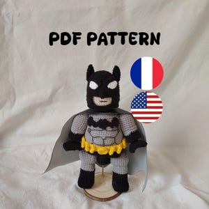 May include: A crochet amigurumi figure of a superhero character with a black cowl, white eyes, a grey torso with black chest emblem, a yellow utility belt, black gloves and boots, and a grey cape. The text PDF PATTERN is visible above.