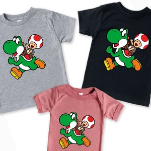 May include: Three shirts featuring a green Yoshi character from the Super Mario Bros. video game series. The Yoshi character is running and carrying a small Toad character on its back. The shirts are in grey, black, and pink.