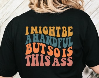 May include: Black t-shirt with the text "I MIGHT BE A HANDFUL BUT SO IS THIS ASS" in retro-style lettering. The text is in shades of gold, teal, orange, and pink. The shirt is a casual wear item.