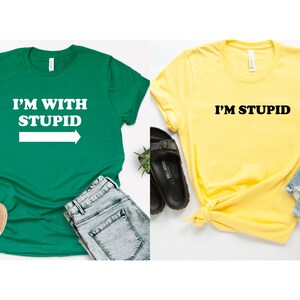 May include: Two t-shirts, one green and one yellow, with a matching design. The green shirt says "I'M WITH STUPID" with an arrow pointing to the right. The yellow shirt says "I'M STUPID".