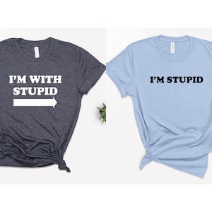 May include: Two t-shirts, one dark blue and one light blue, with a matching couple's design. The dark blue shirt says "I'M WITH STUPID" with a white arrow pointing to the right. The light blue shirt says "I'M STUPID" in black.