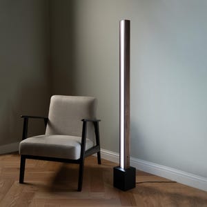 Pu&ograve; includere: Una lampada da terra moderna con una base in legno e una fonte di luce lunga e sottile. La lampada &egrave; accanto a una poltrona beige con gambe nere.